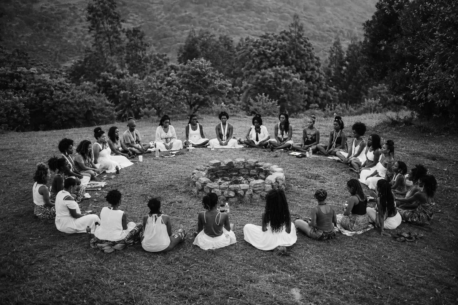Women seated in a wide circle around a stone fire pit in an open landscape